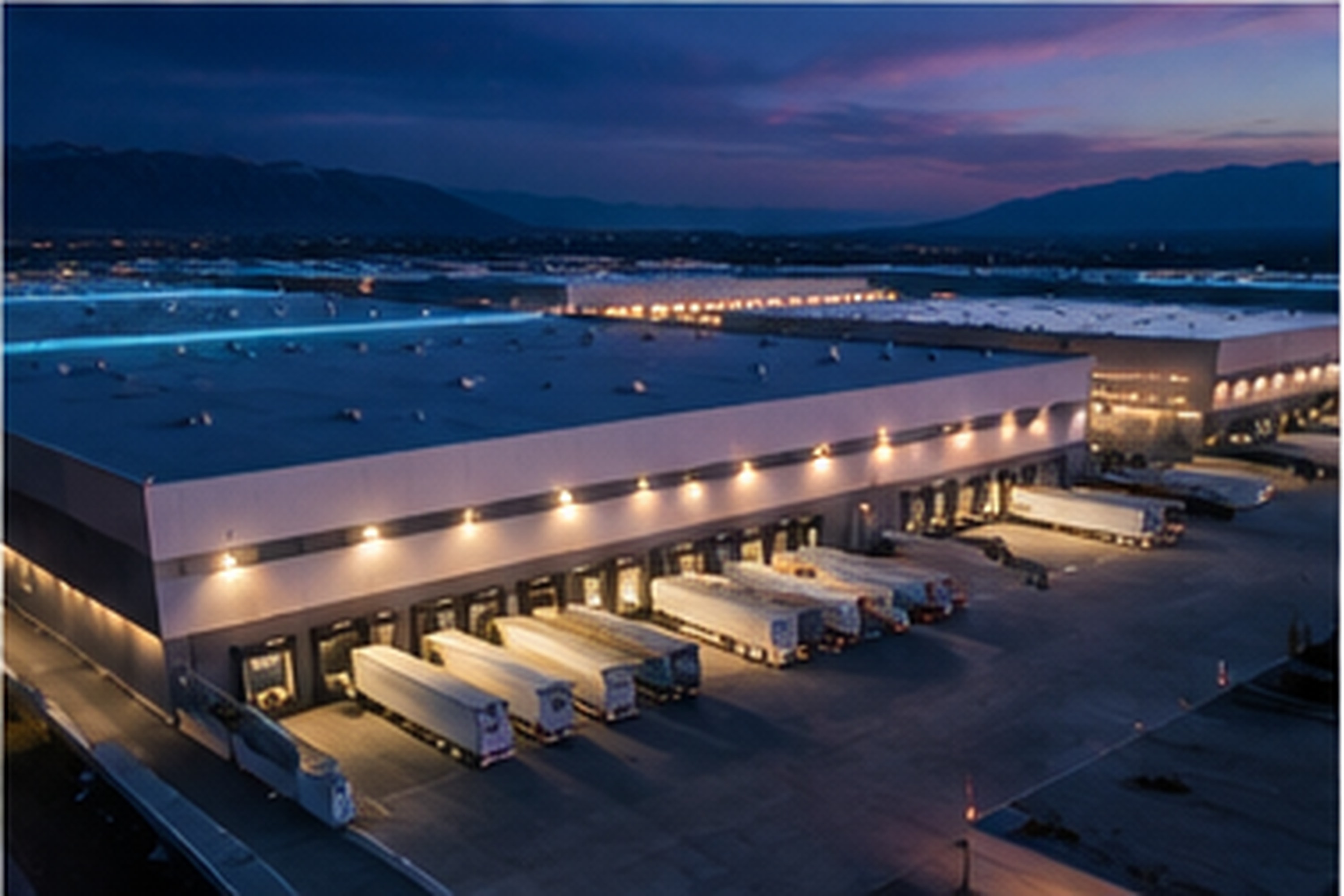 A lit industrial building at twilight with trailers at its loading docks and mountains behind it